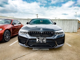 A black BMW car is parked in a parking lot next to a red BMW. The cars are positioned in front of a wooden fence under a partly cloudy sky. The license plate on the black BMW reads 'Texas RIZ-AR'.