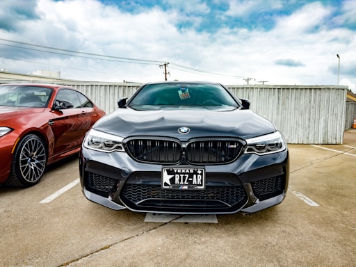 A black BMW car is parked in a parking lot next to a red BMW. The cars are positioned in front of a wooden fence under a partly cloudy sky. The license plate on the black BMW reads 'Texas RIZ-AR'.