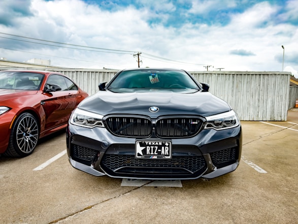 A black BMW car is parked in a parking lot next to a red BMW. The cars are positioned in front of a wooden fence under a partly cloudy sky. The license plate on the black BMW reads 'Texas RIZ-AR'.