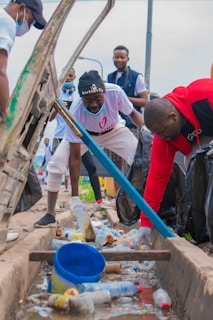 Several individuals are engaged in a community cleanup, collecting trash from a gutter. They are dressed casually and are focused on picking up plastic bottles and litter. The setting is outdoors, with bags to collect the trash.
