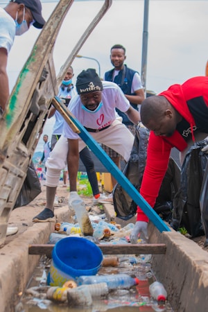 Several individuals are engaged in a community cleanup, collecting trash from a gutter. They are dressed casually and are focused on picking up plastic bottles and litter. The setting is outdoors, with bags to collect the trash.