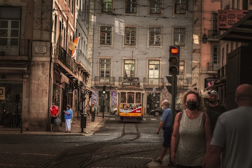 A historic streetcar is positioned on a cobblestone street in the middle of a city intersection. Surrounding the streetcar are old European buildings with balconies and various signs. People are walking and standing near the intersection, wearing casual clothes, with one person wearing a face mask. The traffic light indicates 'BUS'.