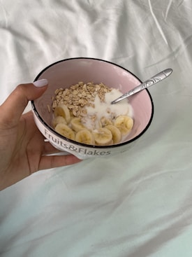 A hand holds a bowl labeled 'Fruits & Flakes' filled with oats, sliced bananas, and milk. A spoon is placed inside the bowl. The background is a soft, light-colored fabric.