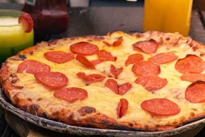 A pepperoni pizza with a crispy crust and melted cheese is placed on a rustic wooden surface. A green drink with a slice of strawberry on the rim and a yellow beverage are visible in the background.