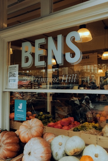 A storefront displays the name 'BENS Primrose Hill' with large glass windows. Inside, there are lamps hanging from the ceiling casting warm light. Outside the window, an array of pumpkins and other fruits are neatly arranged in wooden crates. A blue Deliveroo sign is also visible, indicating delivery availability. A reflection shows a person bundled in winter clothing walking by.