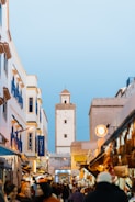 A bustling street scene in a Moroccan city, featuring white and blue buildings with a large tower in the background. The street is filled with people, shops, and colorful market stalls. Various goods are displayed, creating a vibrant and lively atmosphere under a clear blue sky.