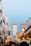 A bustling street scene in a Moroccan city, featuring white and blue buildings with a large tower in the background. The street is filled with people, shops, and colorful market stalls. Various goods are displayed, creating a vibrant and lively atmosphere under a clear blue sky.