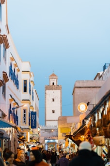 A bustling street scene in a Moroccan city, featuring white and blue buildings with a large tower in the background. The street is filled with people, shops, and colorful market stalls. Various goods are displayed, creating a vibrant and lively atmosphere under a clear blue sky.