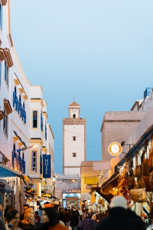 A bustling street scene in a Moroccan city, featuring white and blue buildings with a large tower in the background. The street is filled with people, shops, and colorful market stalls. Various goods are displayed, creating a vibrant and lively atmosphere under a clear blue sky.