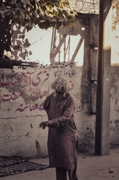 A person wearing a worn-out traditional garment stands on a street. The background features a wall with faded graffiti and exposed brick. Above, some leaves from a tree hang into the frame, casting shadows on the wall.