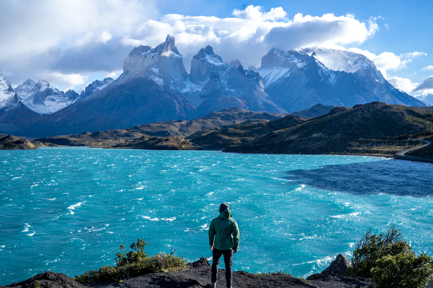 A hiker standing on a rocky mountain overlook gazing at the turquoise glacial lake and the dramatic Patagonian landscape of Torres del Paine National Park in Chile with peaks in the background