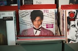Vinyl records are displayed in a record store, with a prominent record at the front featuring a person with a serious expression, dressed in a light purple jacket and white shirt. The texts on the record include phrases like 'Love Thy Neighbor,' 'Annie Christian Sentenced to Die!' and 'Do You Believe in God?'. Other visible records in the background are organized under categories like 'Prince,' 'Pink,' and 'Red Hot Chili Peppers.'