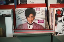 Vinyl records are displayed in a record store, with a prominent record at the front featuring a person with a serious expression, dressed in a light purple jacket and white shirt. The texts on the record include phrases like 'Love Thy Neighbor,' 'Annie Christian Sentenced to Die!' and 'Do You Believe in God?'. Other visible records in the background are organized under categories like 'Prince,' 'Pink,' and 'Red Hot Chili Peppers.'