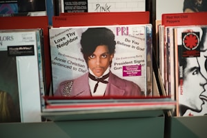 Vinyl records are displayed in a record store, with a prominent record at the front featuring a person with a serious expression, dressed in a light purple jacket and white shirt. The texts on the record include phrases like 'Love Thy Neighbor,' 'Annie Christian Sentenced to Die!' and 'Do You Believe in God?'. Other visible records in the background are organized under categories like 'Prince,' 'Pink,' and 'Red Hot Chili Peppers.'