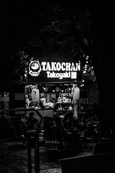 A small outdoor food stall with a sign reading 'Takochan Takoyaki' illuminated in the night. Several people are seated on chairs around the stall, and the area is dimly lit with some decorative lanterns. The ambiance appears cozy and intimate, with shelves displaying various items inside the stall.