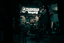 A cheerful food cart with colorful signage and happy customers enjoying their takoyaki outdoors.