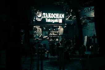 A dimly lit outdoor food stall with a glowing sign reading TAKOCHAN Takoyaki. Several people are gathered around, some seated, while others seem to be ordering. Lanterns and small lights illuminate the area beneath a canopy of trees.