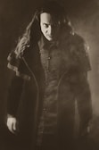 A person with long hair wearing dark, detailed clothing stands in a dramatic pose. The lighting is low and creates a mysterious and intense atmosphere. The image has a sepia tone and a foggy element, adding to the eerie effect.