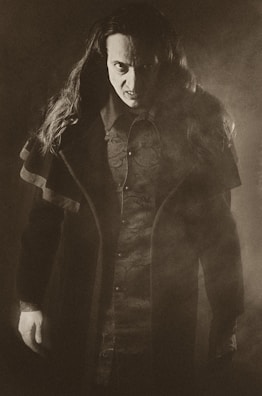 A person with long hair wearing dark, detailed clothing stands in a dramatic pose. The lighting is low and creates a mysterious and intense atmosphere. The image has a sepia tone and a foggy element, adding to the eerie effect.