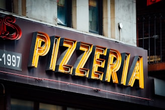A close-up view of an illuminated 'PIZZERIA' sign mounted on the exterior of a building. The letters are large and bold, with a mix of yellow and black colors. Part of a building facade with windows and other sign details can be seen in the background.