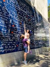 A couple interacts playfully in front of a large blue wall filled with the words 'I love you' in various languages, written in white. The woman, wearing a light-colored dress, points upwards while being lifted by the man, who is in casual attire with a purple shirt and shorts. Shadows of trees extend across the wall and pavement, suggesting a sunny day.