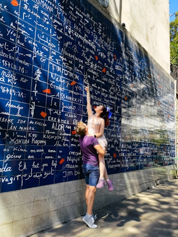 A couple interacts playfully in front of a large blue wall filled with the words 'I love you' in various languages, written in white. The woman, wearing a light-colored dress, points upwards while being lifted by the man, who is in casual attire with a purple shirt and shorts. Shadows of trees extend across the wall and pavement, suggesting a sunny day.