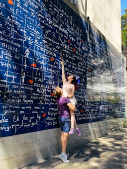 A couple interacts playfully in front of a large blue wall filled with the words 'I love you' in various languages, written in white. The woman, wearing a light-colored dress, points upwards while being lifted by the man, who is in casual attire with a purple shirt and shorts. Shadows of trees extend across the wall and pavement, suggesting a sunny day.