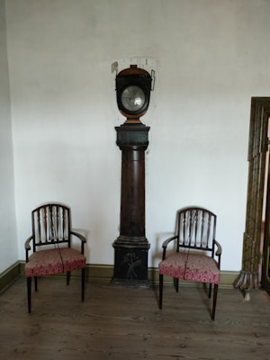 A vintage room features a tall, dark wooden pedestal clock centered between two matching chairs. The chairs have ornate wooden backs and red upholstered seats. The walls are plain and white, while the floor is made of hardwood planks.