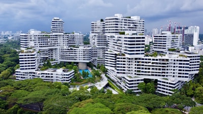 A modern residential complex with white facades and geometric architecture is surrounded by lush greenery. The buildings are stacked and interconnected in an intricate, unique pattern. There are swimming pools, pathways, and walking bridges visible within the complex. The skyline of a city with high-rise buildings can be seen in the background under a cloudy sky.