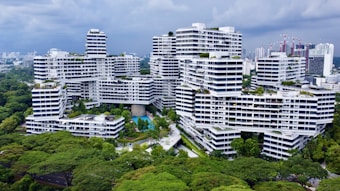 A modern residential complex with white facades and geometric architecture is surrounded by lush greenery. The buildings are stacked and interconnected in an intricate, unique pattern. There are swimming pools, pathways, and walking bridges visible within the complex. The skyline of a city with high-rise buildings can be seen in the background under a cloudy sky.
