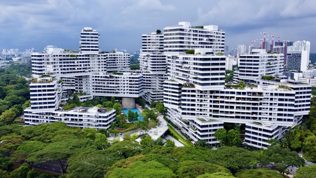 A modern residential complex with white facades and geometric architecture is surrounded by lush greenery. The buildings are stacked and interconnected in an intricate, unique pattern. There are swimming pools, pathways, and walking bridges visible within the complex. The skyline of a city with high-rise buildings can be seen in the background under a cloudy sky.