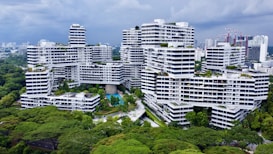 A modern residential complex with white facades and geometric architecture is surrounded by lush greenery. The buildings are stacked and interconnected in an intricate, unique pattern. There are swimming pools, pathways, and walking bridges visible within the complex. The skyline of a city with high-rise buildings can be seen in the background under a cloudy sky.