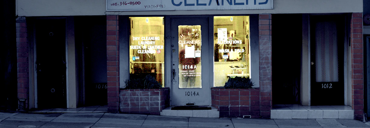 A dry cleaning shop named 'HLP Cleaners' is located on a dimly lit street. The store has a prominent sign above the entrance, and its windows are illuminated with a warm yellow light. A sign on the door lists services such as dry cleaning, laundry, and suede & leather cleaning. The building exterior is simple with a combination of dark tiles and light-colored upper walls. The entrance is flanked by two closed doors, likely of neighboring establishments.