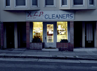 A dry cleaning shop named 'HLP Cleaners' is located on a dimly lit street. The store has a prominent sign above the entrance, and its windows are illuminated with a warm yellow light. A sign on the door lists services such as dry cleaning, laundry, and suede & leather cleaning. The building exterior is simple with a combination of dark tiles and light-colored upper walls. The entrance is flanked by two closed doors, likely of neighboring establishments.