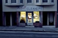 A dry cleaning shop named 'HLP Cleaners' is located on a dimly lit street. The store has a prominent sign above the entrance, and its windows are illuminated with a warm yellow light. A sign on the door lists services such as dry cleaning, laundry, and suede & leather cleaning. The building exterior is simple with a combination of dark tiles and light-colored upper walls. The entrance is flanked by two closed doors, likely of neighboring establishments.
