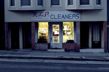 A dry cleaning shop named 'HLP Cleaners' is located on a dimly lit street. The store has a prominent sign above the entrance, and its windows are illuminated with a warm yellow light. A sign on the door lists services such as dry cleaning, laundry, and suede & leather cleaning. The building exterior is simple with a combination of dark tiles and light-colored upper walls. The entrance is flanked by two closed doors, likely of neighboring establishments.