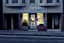 A dry cleaning shop named 'HLP Cleaners' is located on a dimly lit street. The store has a prominent sign above the entrance, and its windows are illuminated with a warm yellow light. A sign on the door lists services such as dry cleaning, laundry, and suede & leather cleaning. The building exterior is simple with a combination of dark tiles and light-colored upper walls. The entrance is flanked by two closed doors, likely of neighboring establishments.