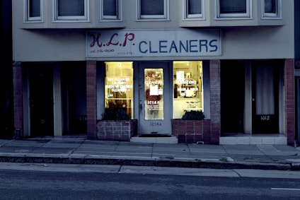 A dry cleaning shop named 'HLP Cleaners' is located on a dimly lit street. The store has a prominent sign above the entrance, and its windows are illuminated with a warm yellow light. A sign on the door lists services such as dry cleaning, laundry, and suede & leather cleaning. The building exterior is simple with a combination of dark tiles and light-colored upper walls. The entrance is flanked by two closed doors, likely of neighboring establishments.