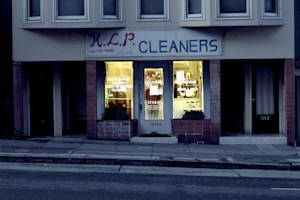 A dry cleaning shop named 'HLP Cleaners' is located on a dimly lit street. The store has a prominent sign above the entrance, and its windows are illuminated with a warm yellow light. A sign on the door lists services such as dry cleaning, laundry, and suede & leather cleaning. The building exterior is simple with a combination of dark tiles and light-colored upper walls. The entrance is flanked by two closed doors, likely of neighboring establishments.
