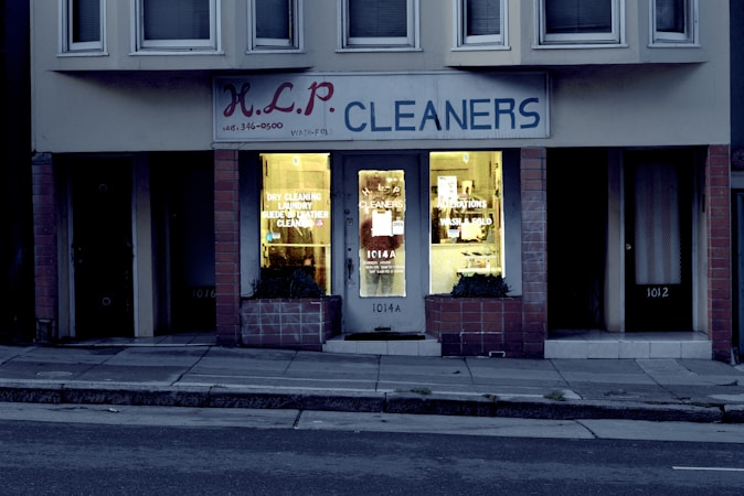 A dry cleaning shop named 'HLP Cleaners' is located on a dimly lit street. The store has a prominent sign above the entrance, and its windows are illuminated with a warm yellow light. A sign on the door lists services such as dry cleaning, laundry, and suede & leather cleaning. The building exterior is simple with a combination of dark tiles and light-colored upper walls. The entrance is flanked by two closed doors, likely of neighboring establishments.