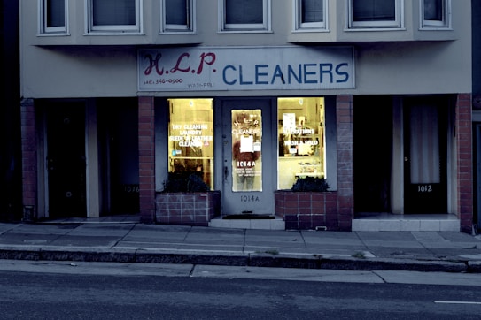 A dry cleaning shop named 'HLP Cleaners' is located on a dimly lit street. The store has a prominent sign above the entrance, and its windows are illuminated with a warm yellow light. A sign on the door lists services such as dry cleaning, laundry, and suede & leather cleaning. The building exterior is simple with a combination of dark tiles and light-colored upper walls. The entrance is flanked by two closed doors, likely of neighboring establishments.