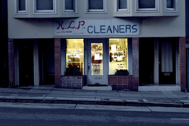 A dry cleaning shop named 'HLP Cleaners' is located on a dimly lit street. The store has a prominent sign above the entrance, and its windows are illuminated with a warm yellow light. A sign on the door lists services such as dry cleaning, laundry, and suede & leather cleaning. The building exterior is simple with a combination of dark tiles and light-colored upper walls. The entrance is flanked by two closed doors, likely of neighboring establishments.