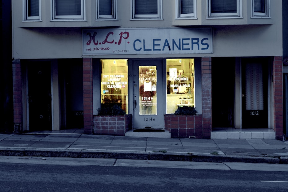 A dry cleaning shop named 'HLP Cleaners' is located on a dimly lit street. The store has a prominent sign above the entrance, and its windows are illuminated with a warm yellow light. A sign on the door lists services such as dry cleaning, laundry, and suede & leather cleaning. The building exterior is simple with a combination of dark tiles and light-colored upper walls. The entrance is flanked by two closed doors, likely of neighboring establishments.