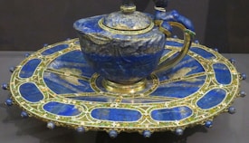An intricately designed teapot and saucer made from a deep blue stone, possibly lapis lazuli, featuring gold accents and detailed craftsmanship. The teapot has a sturdy handle and a domed lid with elegant embellishments. The accompanying saucer is large and ornately decorated with matching blue stone inlays bordered by gold.