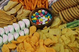 An assortment of snacks and treats arranged on a platter, featuring colorful candy-coated chocolates in a wooden bowl at the center. Surrounding these are various types of crispy snacks including potato chips, fish-shaped crackers, pretzel sticks, and spiced snack sticks. Additionally, there are sliced sweets with a layered appearance and cookies piled around the perimeter.