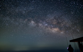 A vast, star-filled night sky showcases the Milky Way galaxy in stunning detail. A person is silhouetted in the foreground, standing next to a structure with antenna-like objects.
