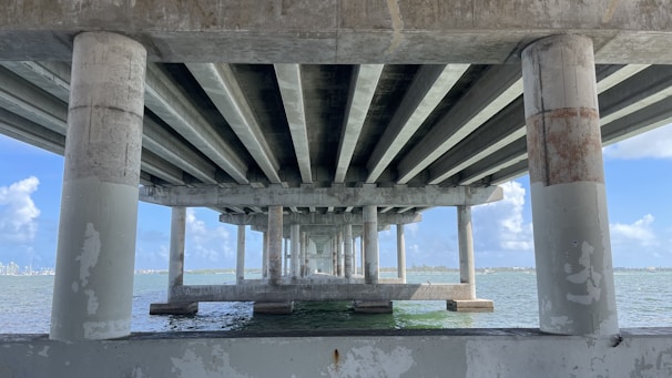 Concrete bridge supports and beams span across the water, creating a series of repeating geometric patterns. The blue sky and distant horizon are visible between the columns, with sunlight illuminating portions of the structure and the water below.