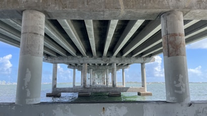 Concrete bridge supports and beams span across the water, creating a series of repeating geometric patterns. The blue sky and distant horizon are visible between the columns, with sunlight illuminating portions of the structure and the water below.