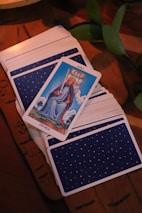 A tarot card deck is spread on a wooden table with a single card, the King of Swords, prominently displayed on top. The card shows a king sitting on a throne holding a sword. The deck's back design features small yellow stars on a blue background. Green leaves are partially visible at the side.