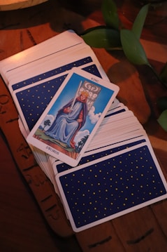 A tarot card deck is spread on a wooden table with a single card, the King of Swords, prominently displayed on top. The card shows a king sitting on a throne holding a sword. The deck's back design features small yellow stars on a blue background. Green leaves are partially visible at the side.